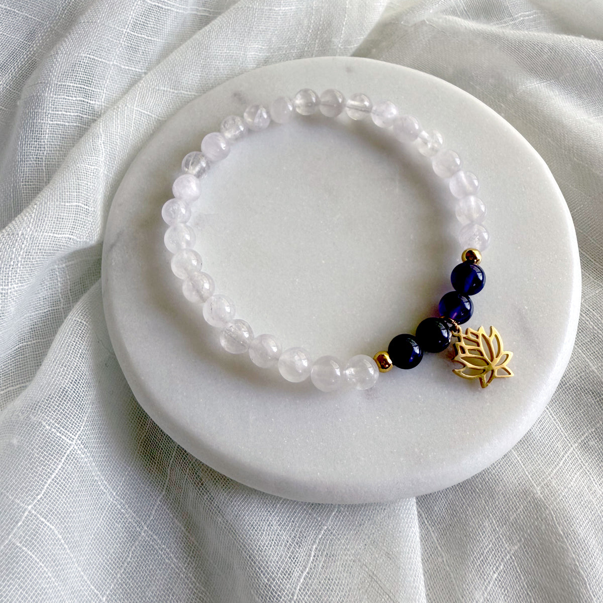 “See Truth Within” bracelet by Moksha Jewelry, featuring amethyst and lavender amethyst beads with clear quartz accents and a gold-plated lotus charm, displayed on a round white marble plate with soft fabric—crafted to support third eye chakra activation, intuition, and inner clarity.