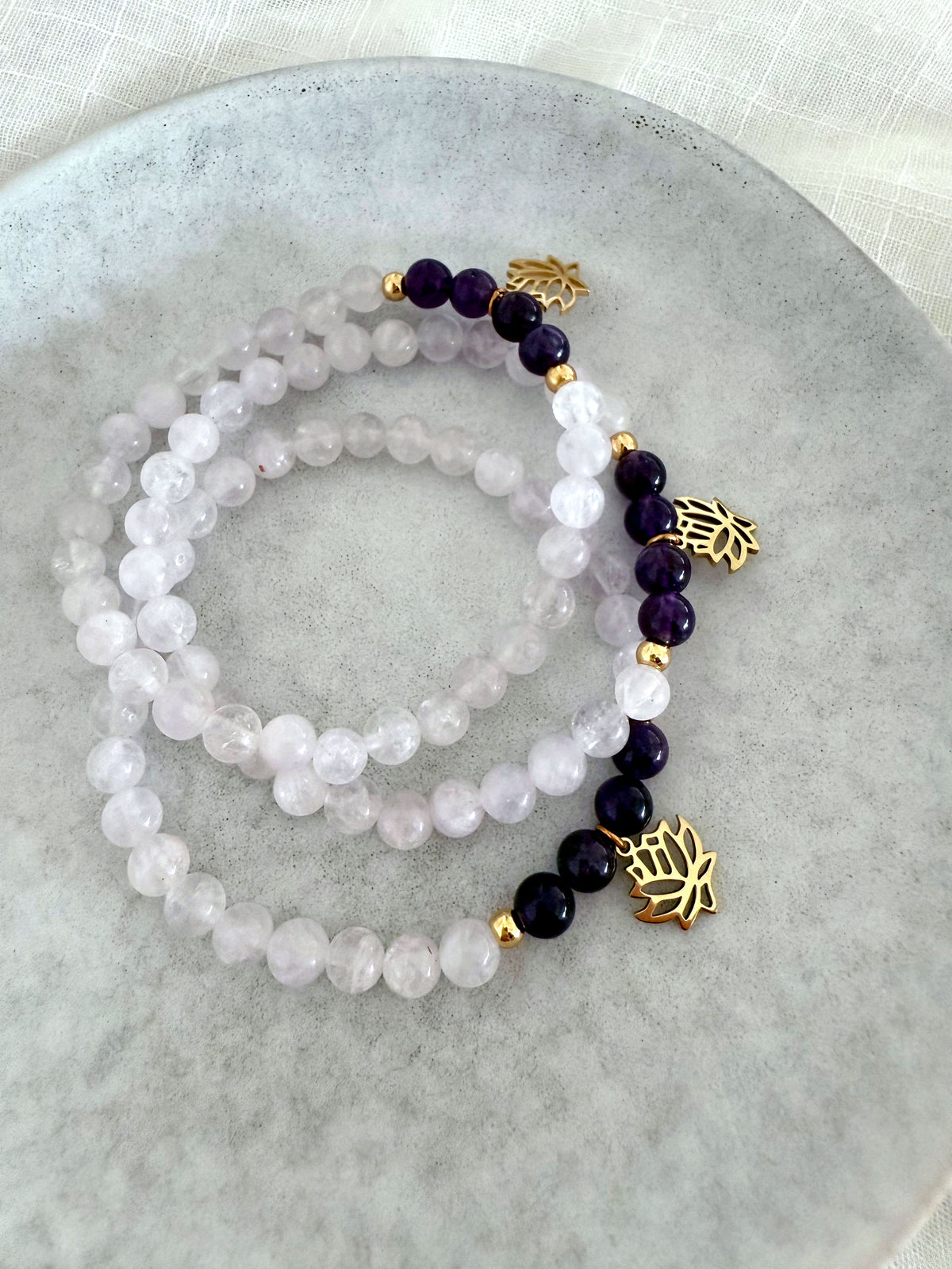 Set of “See Truth Within” bracelets by Moksha Jewelry, featuring lavender amethyst and deep amethyst beads with gold lotus charms, elegantly displayed on a gray ceramic plate with soft fabric—designed to activate the third eye chakra and enhance intuition, clarity, and inner wisdom.
