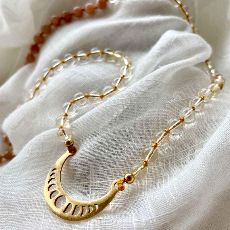Close-up of a Mala made of Citrine, peach Moonstone and Aquamarine beads with a gold plated Crescent Moon pendant on a white light fabric.