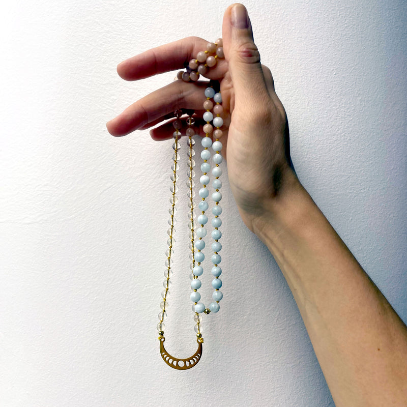 A hand holds a Mala made of Citrine, peach Moonstone and Aquamarine beads with a gold plated Crescent Moon pendant.
