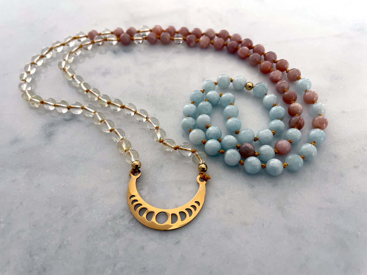 A Mala made of Citrine, peach Moonstone and Aquamarine beads with a gold plated Crescent Moon pendant on a Marble countertop.
