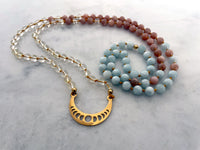 A Mala made of Citrine, peach Moonstone and Aquamarine beads with a gold plated Crescent Moon pendant on a Marble countertop.
