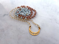 A Mala made of Citrine, peach Moonstone and Aquamarine beads with a gold plated Crescent Moon pendant on a Marble countertop.