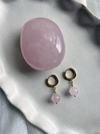 A pair of gold plated  huggie hoops with a 10mm round Rose Quartz bead next to a Rose Quartz gemstone on a gray ceramic plate.