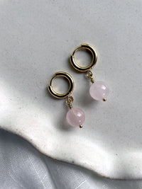 A pair of gold plated  huggie hoops with a 10mm round Rose Quartz bead on a gray ceramic plate.