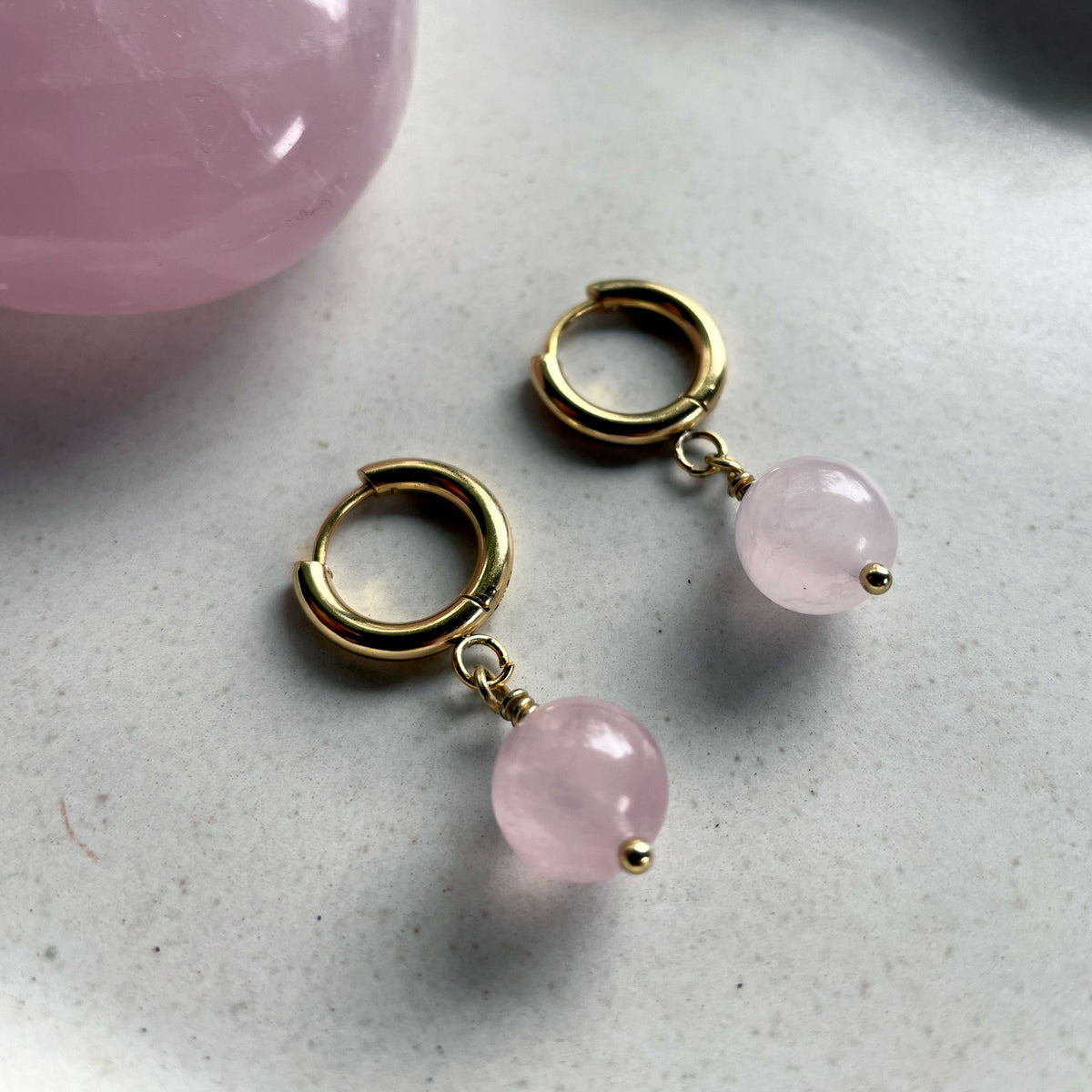 A close-up of a pair of gold plated  huggie hoops with a 10mm round Rose Quartz bead next to a Rose Quartz gemstone on a gray ceramic plate.