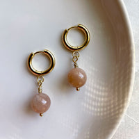 A close-up of a pair of gold plated  huggie hoops with a 10mm round Peach Moonstone  bead on a white ceramic plate.