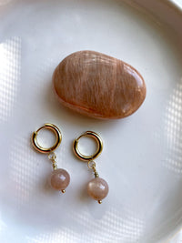 A close-up of a pair of gold plated  huggie hoops with a 10mm round Peach Moonstone  bead on a white ceramic plate next to a peach Moonstone gemstone.