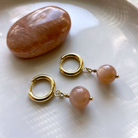 A close-up of a pair of gold plated  huggie hoops with a 10mm round Peach Moonstone  bead on a white ceramic plate next to a peach Moonstone gemstone.