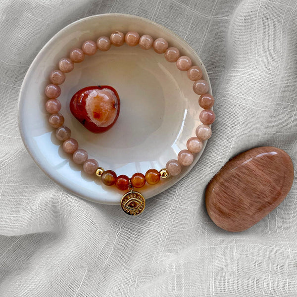 A bracelet made of peach moonstone beads, 4 carnelian beads and 2 small golden beads with a golden evil eye charm in the middle on a ceramic plate. In the middle of the plate is a carnelian gemstone. Next to the plate is a moonstone. All items are on a light fabric.