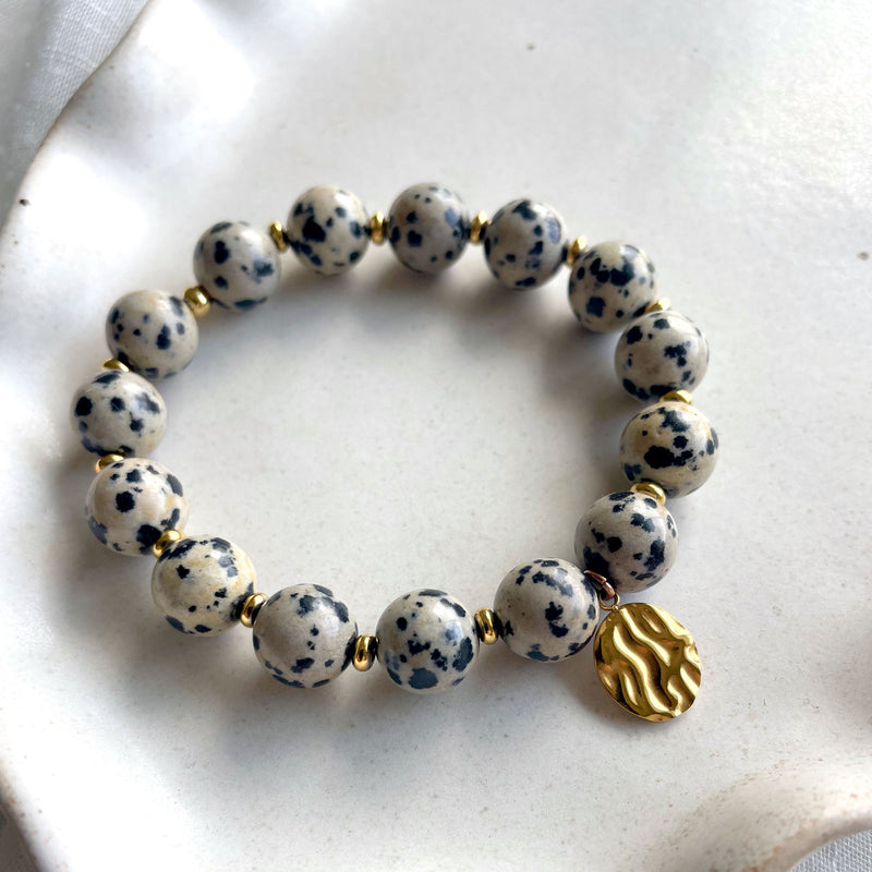 A Dalmatian Jasper beaded bracelet with gold spacer beads and a textured gold charm, elegantly displayed on a white ceramic plate. The speckled black and beige gemstones create a striking contrast, while the gold accents add a touch of sophistication.