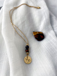A fine necklace with a gold plated Capricorn Zodiac charm, a Smoky Quartz and a Tiger Eye bead on a fine gold plated chain, laying on a light fabric. Next to the necklace is a tiger eye gemstone.