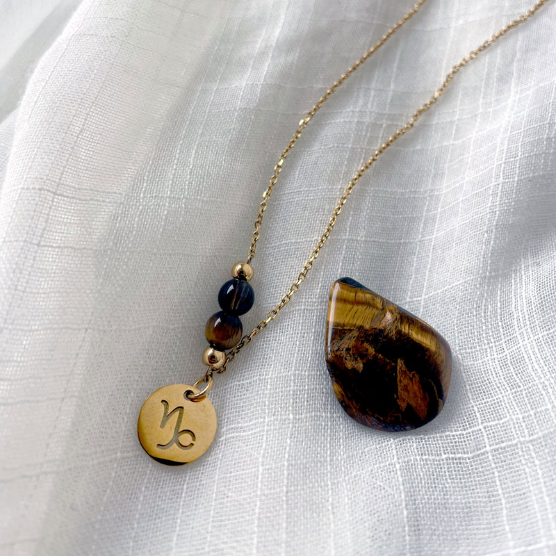 A close-up of a fine necklace with a gold plated Capricorn Zodiac charm, a Smoky Quartz and a Tiger Eye bead on a fine gold plated chain, laying on a light fabric. Next to the necklace is a tiger eye gemstone.