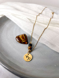 A close-up of a fine necklace with a gold plated Capricorn Zodiac charm, a Smoky Quartz and a Tiger Eye bead on a fine gold plated chain, laying on a ceramic plate. Next to the necklace is a tiger eye gemstone.