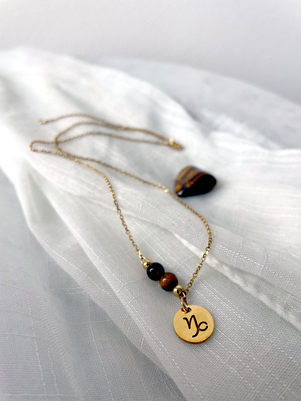 A close-up of a fine necklace with a gold plated Capricorn Zodiac charm, a Smoky Quartz and a Tiger Eye bead on a fine gold plated chain, laying on a light fabric. Next to the necklace is a tiger eye gemstone.