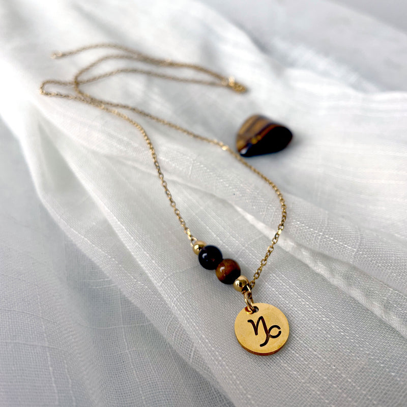 A close-up of a fine necklace with a gold plated Capricorn Zodiac charm, a Smoky Quartz and a Tiger Eye bead on a fine gold plated chain, laying on a light fabric.
