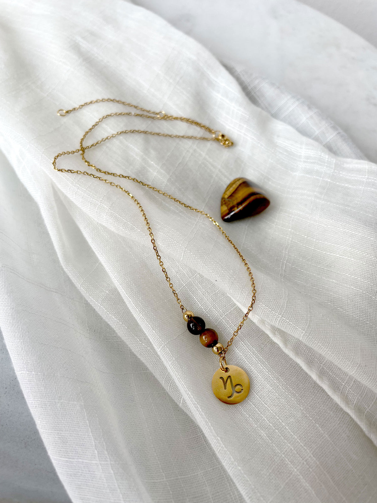 A fine necklace with a gold plated Capricorn Zodiac charm, a Smoky Quartz and a Tiger Eye bead on a fine gold plated chain, laying on a light fabric. Next to the necklace is a tiger eye gemstone.