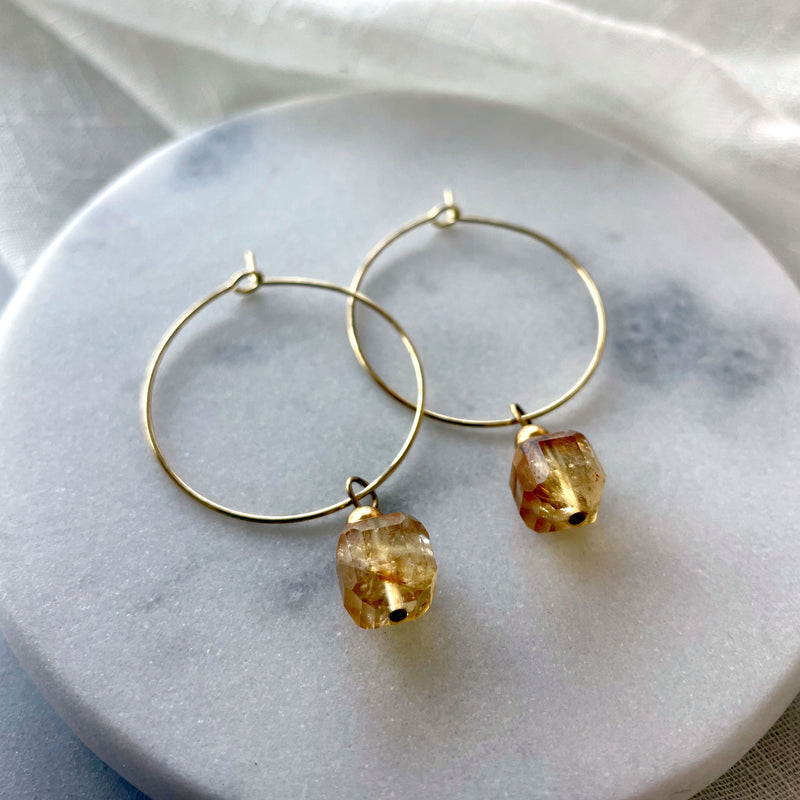 Close-up of Earrings with a 8.5mm faceted Citrine cube bead and a 4mm gold plated bead on a 30mm diameter fine gold plated hoop on a white marble plate.