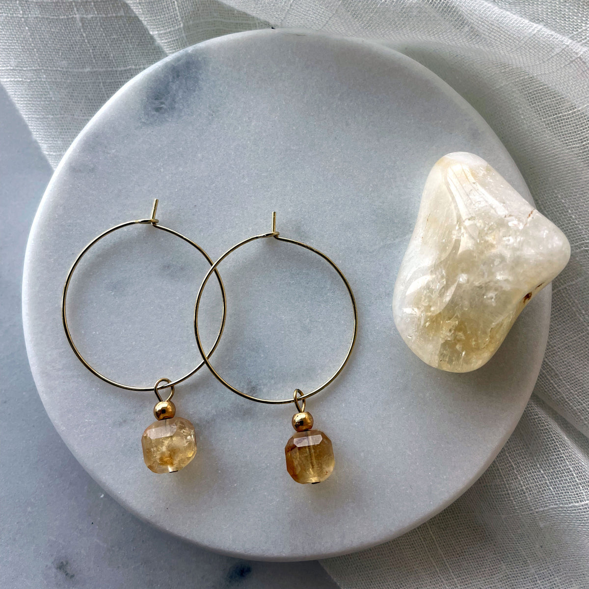 Earrings with a 8.5mm faceted Citrine cube bead and a 4mm gold plated bead on a 30mm diameter fine gold plated hoop next to a citrine gemstone on a white marble plate.