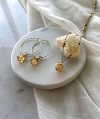 Earrings with a 8.5mm faceted Citrine cube bead and a 4mm gold plated bead on a 30mm diameter fine gold plated hoop next to a fine gold plated necklace with a 10mm round citrine bead lying over a Citrine gemstone on a white marble plate.
