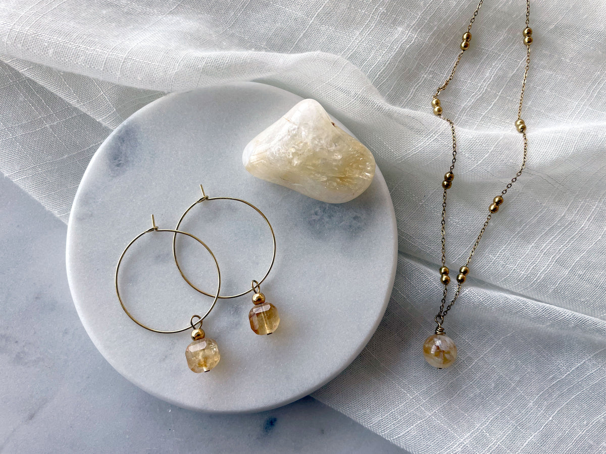 Earrings with a 8.5mm faceted Citrine cube bead and a 4mm gold plated bead on a 30mm diameter fine gold plated hoop next to a Citrine gemstone on a white marble plate. On the right side of the marble plate is a fine gold plated necklace with a 10mm round Citrine bead on a white linen fabric.
