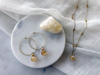 Earrings with a 8.5mm faceted Citrine cube bead and a 4mm gold plated bead on a 30mm diameter fine gold plated hoop next to a Citrine gemstone on a white marble plate. On the right side of the marble plate is a fine gold plated necklace with a 10mm round Citrine bead on a white linen fabric.