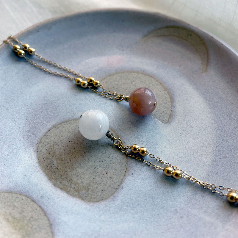 A close-up of two gold plated necklace with a 10mm peach Moonstone bead and a blue Moonstone bead on a delicate chain with 3mm Ball and 1mm Curb Chain on a ceramic plate.