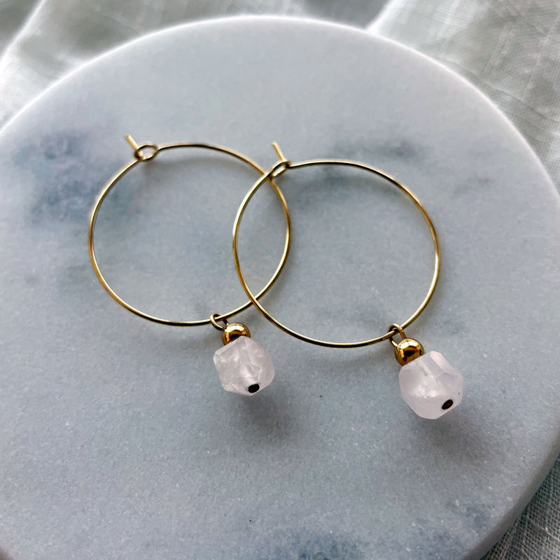 Earrings with a 7mm faceted Rose Quartz cube bead and a 4mm gold plated bead on a 30mm diameter fine gold plated hoop on a white marble plate.