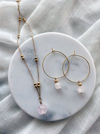 Earrings with a 7mm faceted Rose Quartz cube bead and a 4mm gold plated bead on a 30mm diameter fine gold plated hoop next to a fine gold plated necklace with a 10mm round rose quartz bead lying on a white marble plate.
