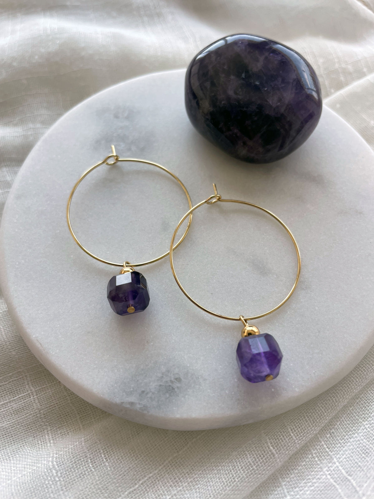 Earrings with a 8.5mm faceted Amethyst cube bead and a 4mm gold plated bead on a 30mm diameter fine gold plated hoop next to an Amethyst gemstone on a white marble plate.