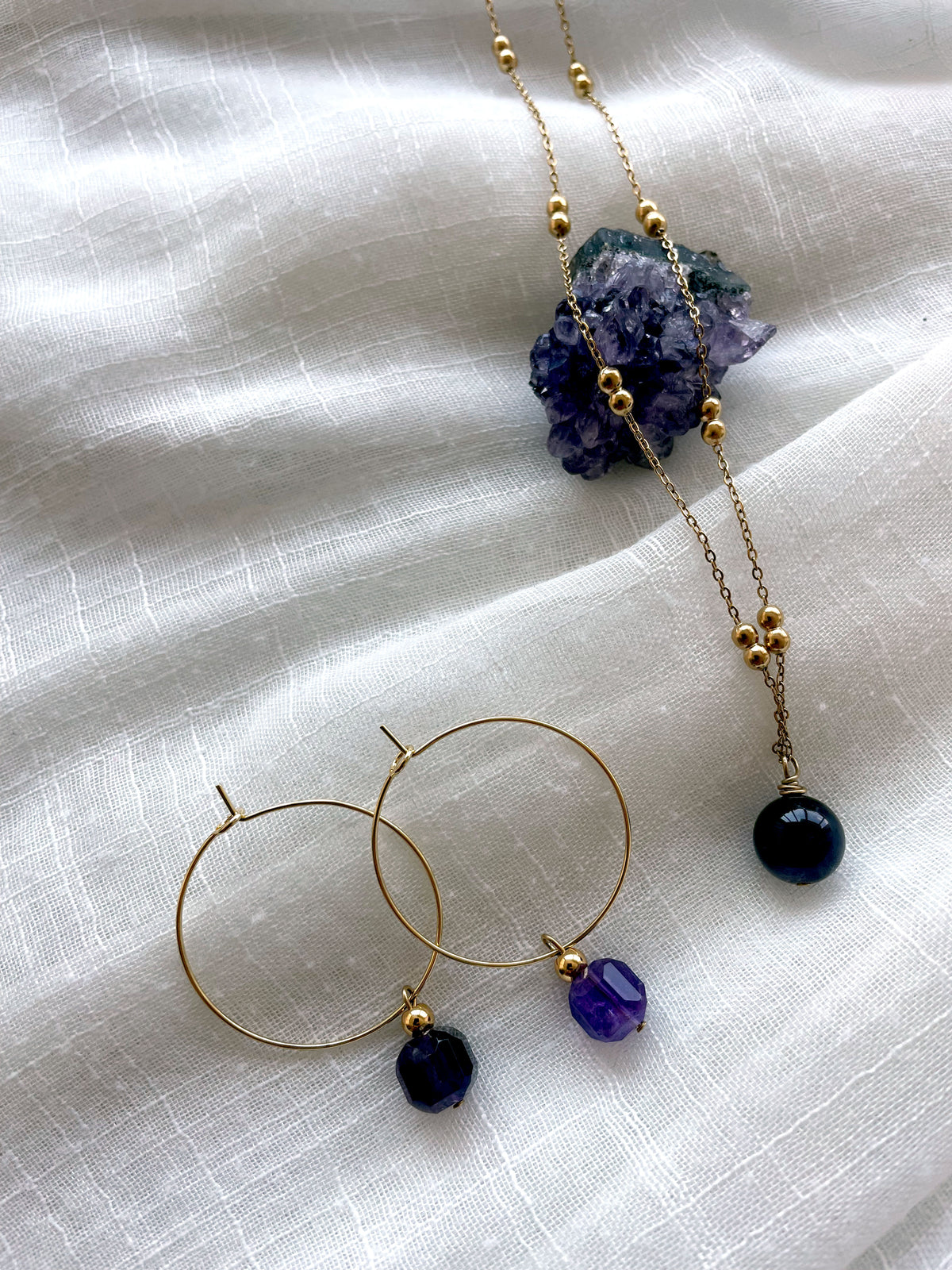 Earrings with a 8.5mm faceted Amethyst cube bead and a 4mm gold plated bead on a 30mm diameter fine gold plated hoop next to a fine gold plated necklace with a 10mm round Amethyst bead lying over a raw Amethyst gemstone on a white linen fabric.